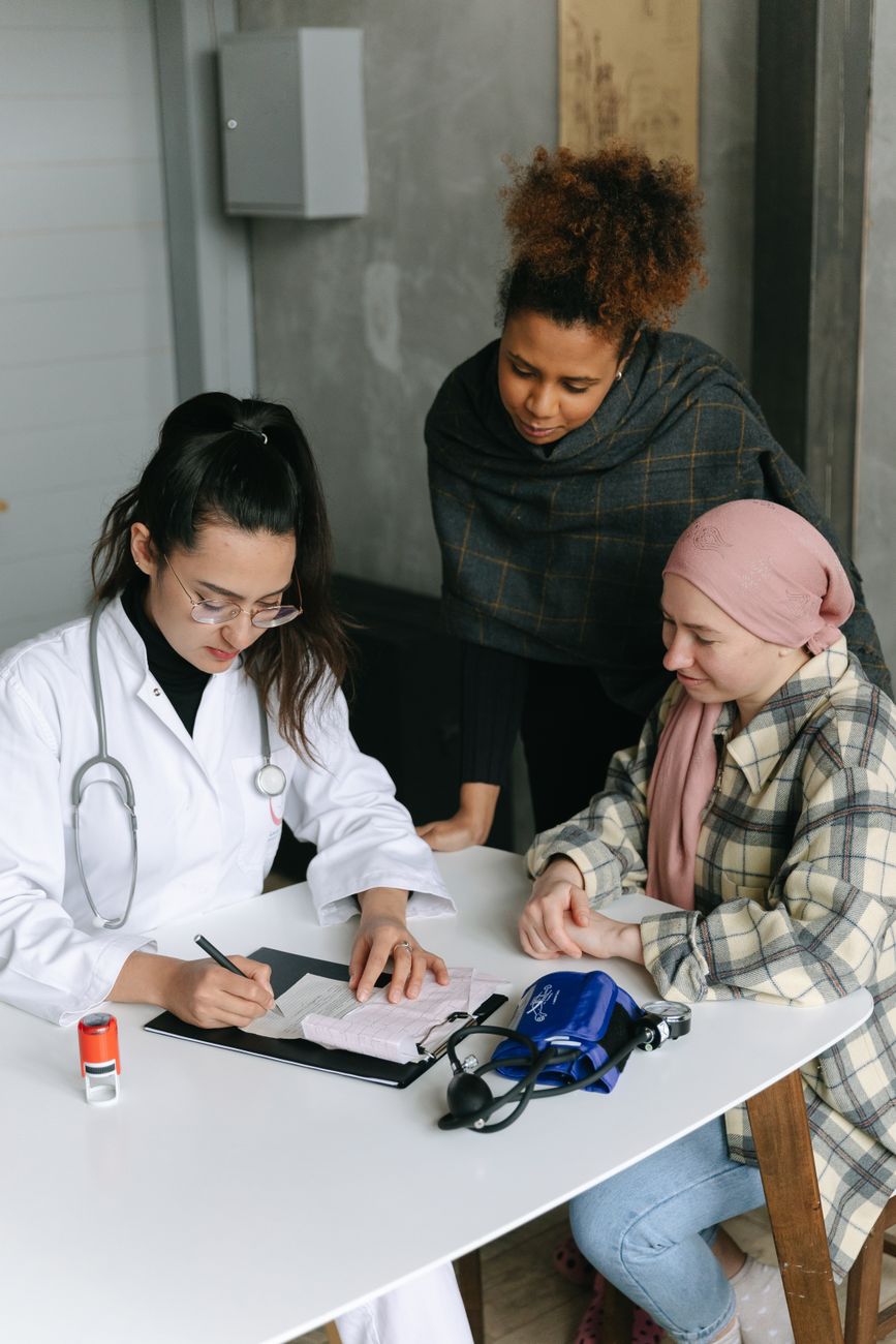 a woman with stethoscope writing on paper beside a woman with headscarf